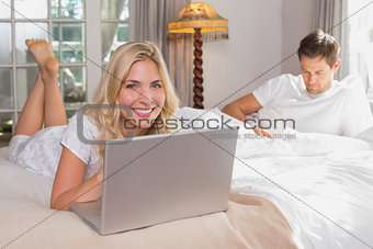 Woman using laptop while man reading book in bed