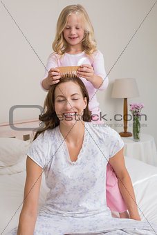 Cute girl combing happy mother's hair in bedroom
