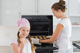 Girl gesturing thumbs up while mother removes cookies from oven