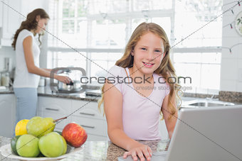 Daughter using laptop with mother cooking food in background at kitchen