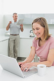 Woman using laptop while man with coffee cup and newspaper in kitchen