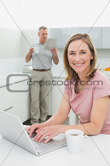 Woman using laptop while man with coffee cup and newspaper in kitchen