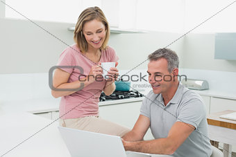 Couple using laptop while woman drinking coffee in kitchen