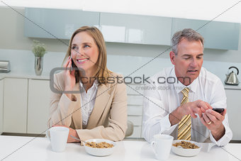 Business couple using cellphones while having breakfast in kitchen