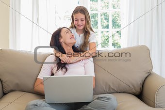 Little girl embracing mother using laptop on sofa
