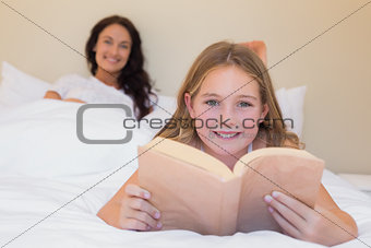 Girl holding book in bed with mother in background