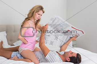 Woman sitting on man as he reads newspaper in bed