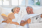 Cheerful mature couple with newspaper and book in bed