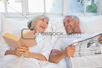 Cheerful mature couple with newspaper and book in bed