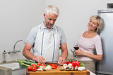 Man chopping vegetables while woman with wine glasses in kitchen