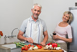 Mature man chopping vegetables while woman with wine glasses in kitchen