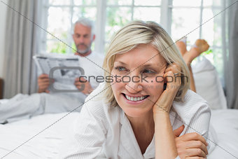 Close-up of a smiling woman with man reading newspaper in background on bed