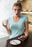 Shocked woman with coffee cup reading text message coffee shop