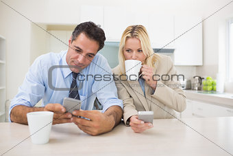Well dressed couple with coffee cups text messaging in the kitchen