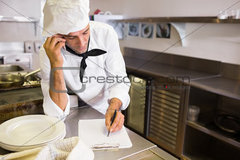 Cook writing on clipboard while using cellphone in kitchen