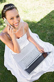 Happy bride with laptop using cellphone on grass