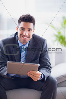Businessman sitting on sofa using his tablet pc smiling at camera