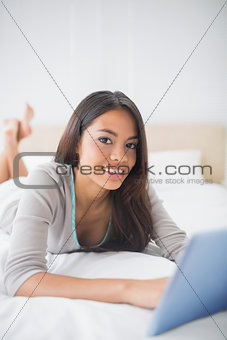 Pretty girl lying on bed using her tablet smiling at camera