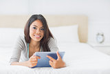 Young pretty girl lying on her bed using her tablet smiling at camera