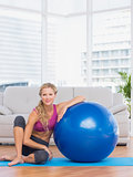 Slim blonde sitting beside exercise ball smiling at camera