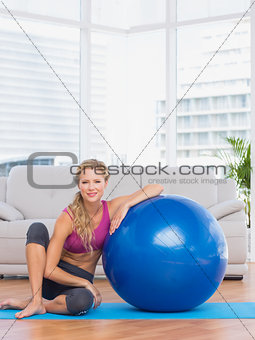 Slim blonde sitting beside exercise ball smiling at camera
