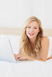 Smiling young blonde lying on her bed using laptop