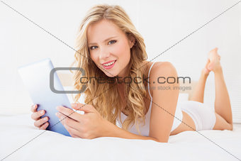 Pretty young blonde smiling at camera on bed using tablet