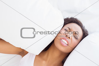 Stressed brunette sitting on bed covering ears with pillow