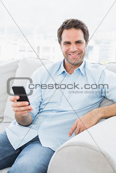 Smiling man sitting on the couch sending a text with smartphone