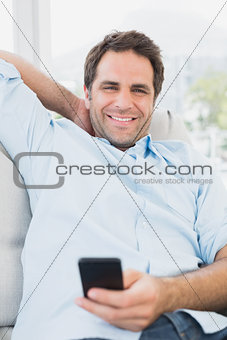 Smiling man sitting on the sofa texting on his phone looking at camera