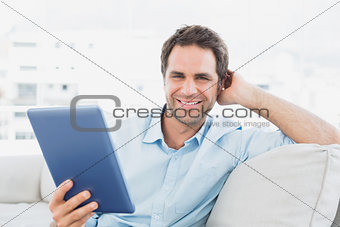 Handsome man sitting on the sofa using his tablet pc smiling at camera