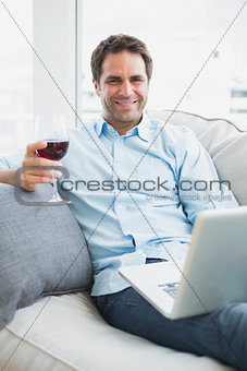 Handsome man relaxing on sofa with glass of red wine using laptop