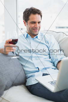 Happy man relaxing on sofa with glass of red wine using laptop