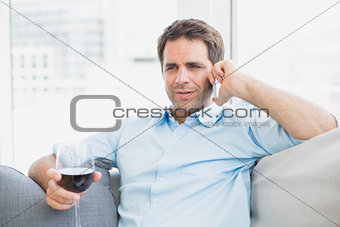 Cheerful man relaxing on sofa with glass of red wine talking on phone