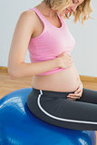 Happy blonde pregnant woman touching belly on exercise ball