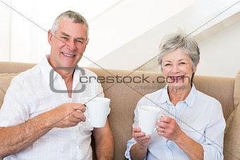Senior couple sitting on couch drinking coffee smiling at camera