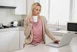 Woman using laptop while drinking coffee in kitchen