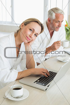 Couple having coffee at breakfast in bathrobes using laptop