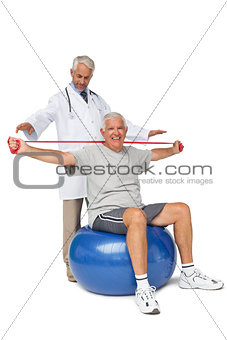Mhysiotherapist looking at senior man sit on exercise ball with yoga belt