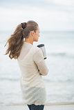 Young woman standing on cold beach with cup of hot beverage. rea
