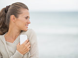 Young woman with cell phone standing on cold beach and looking o