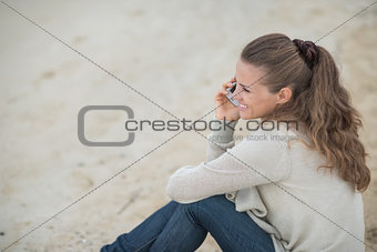 Happy young woman sitting on cold beach and talking mobile phone