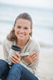 Happy woman sitting on cold beach with cup of hot beverage