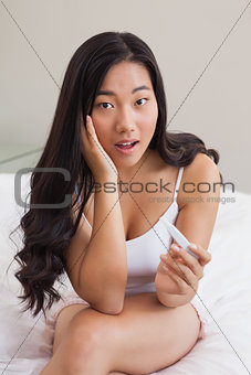 Shocked woman sitting on bed holding pregnancy test
