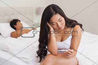 Woman sitting on bed holding pregnancy test as boyfriend sleeps
