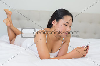 Smiling asian woman lying on bed texting on phone