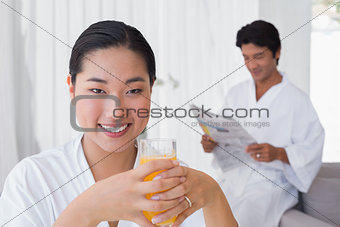 Woman in bathrobe having orange juice with boyfriend in background