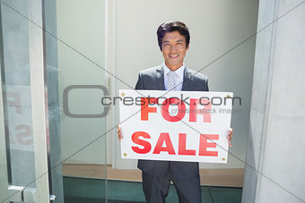 Confident estate agent standing at front door showing for sale sign