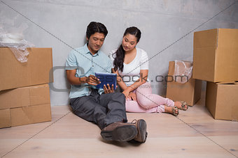 Happy couple sitting on floor using tablet surrounded by boxes