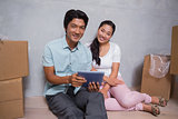 Happy couple sitting on floor using tablet surrounded by boxes